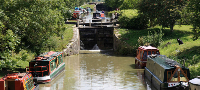 Several colorful canal boats are moored along a narrow, tree-lined canal near a lock gate, with lush greenery surrounding the water and a few boats waiting to pass through the lock.