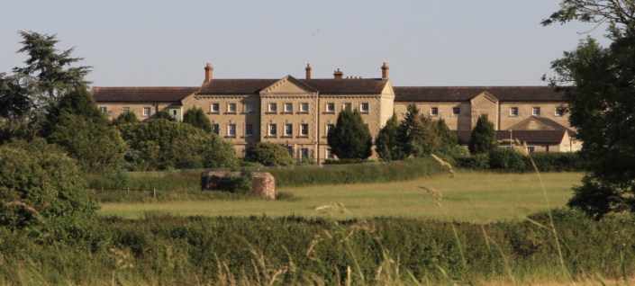 A large, historic stone building with multiple chimneys stands amid green fields and trees under a clear sky, viewed from a distance.