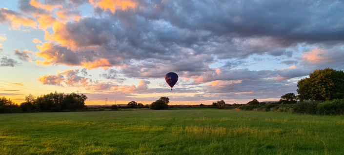 A colorful hot air balloon floats in a partly cloudy sky during sunset over a green grassy field with scattered trees and golden light on the clouds.