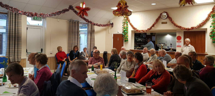 A group of people sit around tables in a festively decorated room, with tinsel and ornaments hanging from the ceiling. They appear to be enjoying a gathering or party, engaged in conversation and dining together.