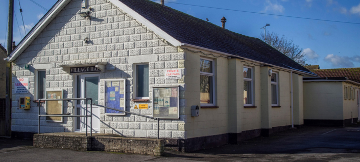 A single-story, cream-colored village hall with a ramp and steps leading to the entrance. Notice boards are mounted on the wall, and signs indicate a private car park. The sky is blue with a few clouds.