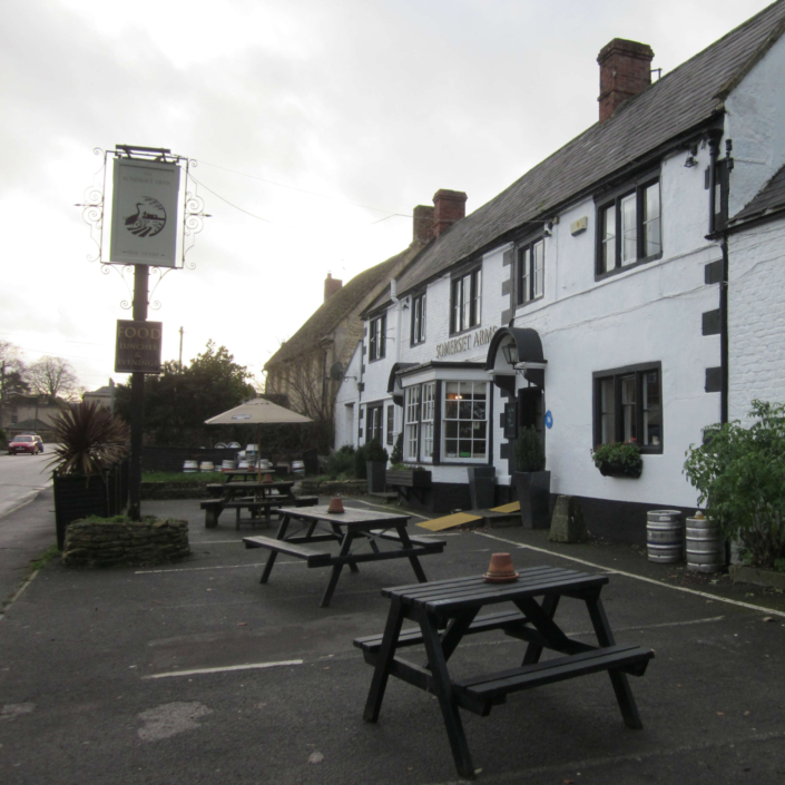 A white, traditional English pub with a sign reading "Somerset Arms," outdoor seating with wooden benches and tables, and a few potted plants on a cloudy day.