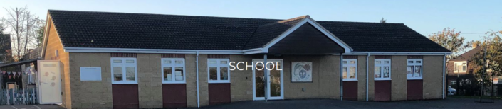 A single-story school building with tan brick walls, white-framed windows, and a dark tiled roof. The word "SCHOOL" is centered over the entrance. Trees and houses are visible in the background.