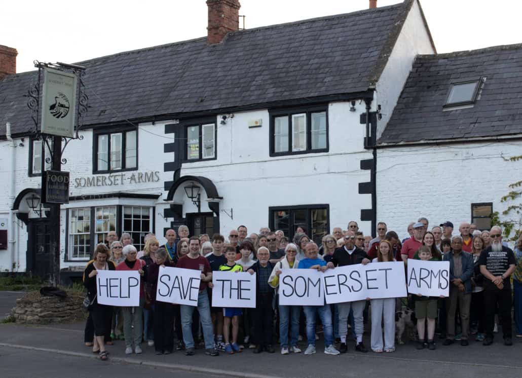A large group of people stands in front of the Somerset Arms pub, holding signs that together read "HELP SAVE THE SOMERSET ARMS." The pub is white with black trim and has a traditional appearance.